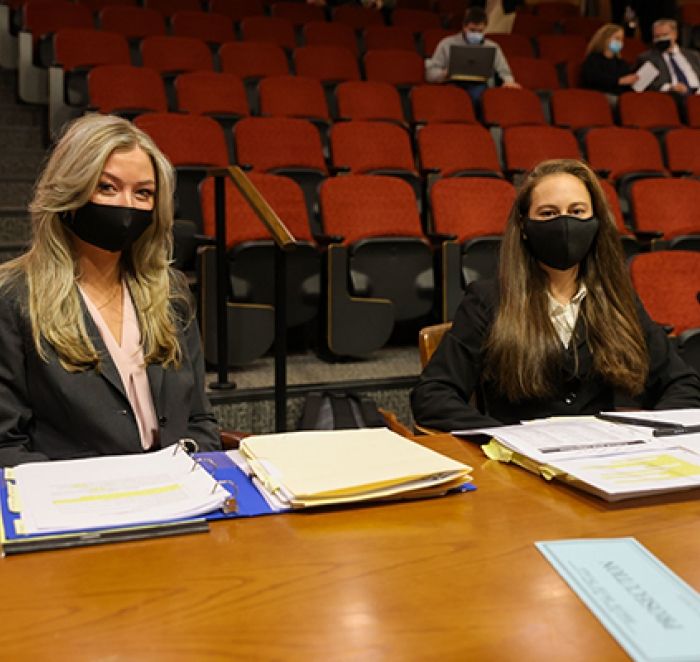 Two female students sit at a large desk facing the camera. They are wearking masks.